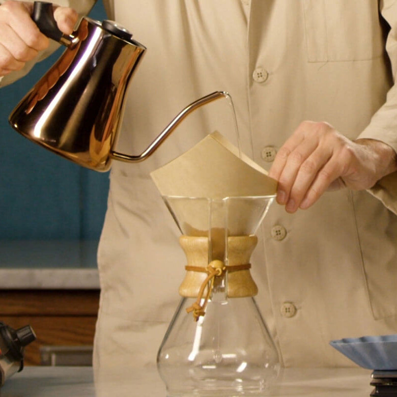 Person making coffee using a pour-over method with a clear glass carafe and metal kettle.